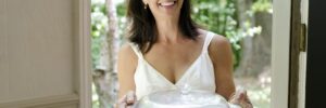 A woman standing at an open front door smiling holding a lidded pot, symbolizing bringing food to a party as a guest