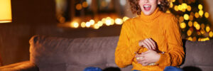 A woman in a sweater sitting on the couch eating a snack and watching something on TV- there are blurry string lights behind her, signifying a warm and cozy night in
