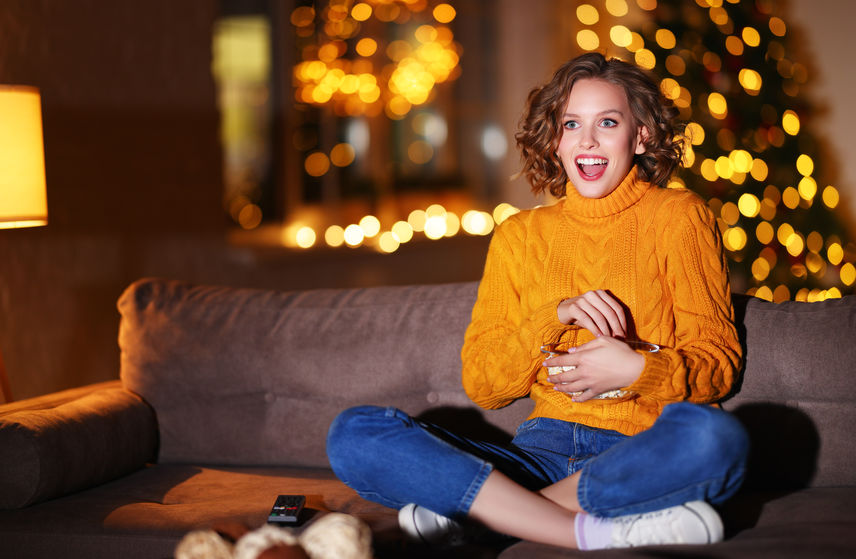 A woman in a sweater sitting on the couch eating a snack and watching something on TV- there are blurry string lights behind her, signifying a warm and cozy night in