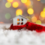 A small wooden house on a white rug wrapped in a red scarf, signifying a cozy home for winter